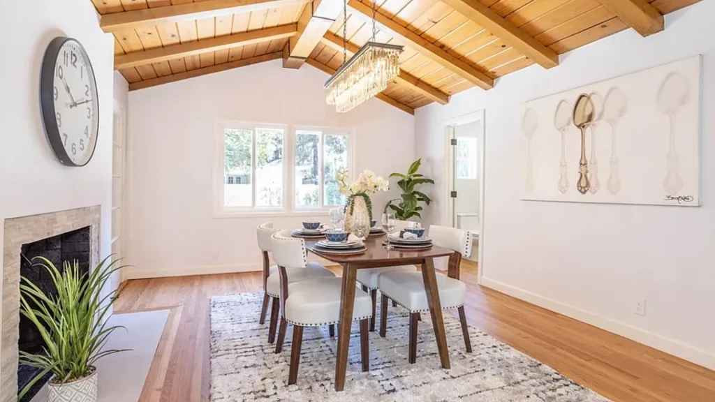 Dining area with a wood table, white chairs and a wall clock under a beamed ceiling